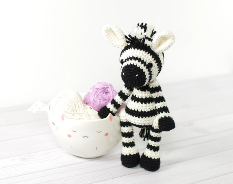 K&ouml;nnte beinhalten: Ein gestricktes Zebra-Spielzeug mit schwarzen und wei&szlig;en Streifen vor einem wei&szlig;en Hintergrund. Das Zebra steht auf einer wei&szlig;en Oberfl&auml;che mit einer wei&szlig;en Keramikschale gef&uuml;llt mit Garn.
