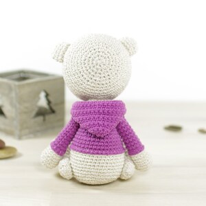 May include: A white crocheted stuffed animal wearing a pink hooded sweater. The animal is sitting on a light wood surface.