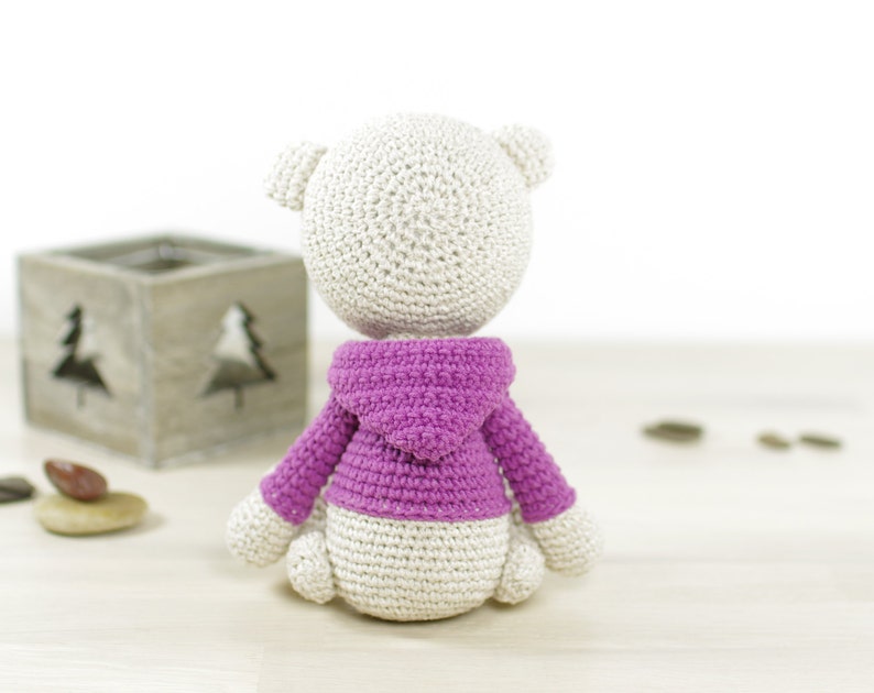 May include: A white crocheted stuffed animal wearing a pink hooded sweater. The animal is sitting on a light wood surface.