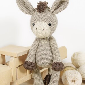 May include: A crocheted donkey toy featuring a light beige body, a cream-coloured muzzle, dark brown hooves, and a dark brown tuft of hair on its head. The donkey has large, upright ears and small black bead eyes, crafted with a textured yarn stitch.