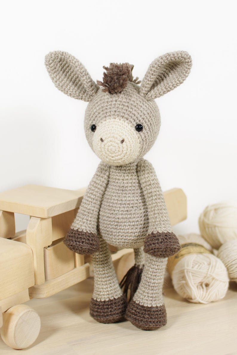 May include: A crocheted donkey toy featuring a light beige body, a cream-coloured muzzle, dark brown hooves, and a dark brown tuft of hair on its head. The donkey has large, upright ears and small black bead eyes, crafted with a textured yarn stitch.