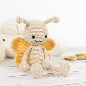 May include: A crocheted beige butterfly doll with yellow wings, black eyes, and a smiling mouth. The handmade toy is sitting on a white wooden surface, with yarn and buttons in the background. A charming, handcrafted item.