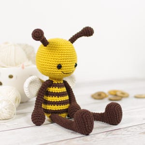 May include: A handmade crochet bee doll with a yellow head and body, brown stripes, and brown limbs. The bee has small black eyes, white wings, and brown antennae. The doll is sitting on a light wood surface.