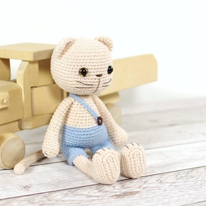 May include: A crocheted cream-colored cat wearing blue overalls sits next to a wooden toy truck. The cat has brown eyes and a stitched-on nose and mouth.