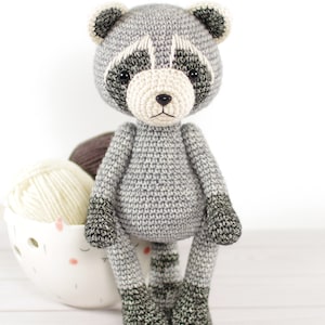 May include: A crocheted raccoon plush toy with gray fur, black eyes, and a white nose. The toy is sitting on a white surface with yarn in the background.