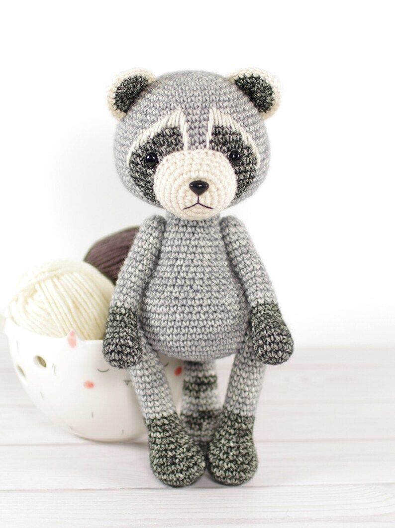 May include: A crocheted raccoon plush toy with gray fur, black eyes, and a white nose. The toy is sitting on a white surface with yarn in the background.