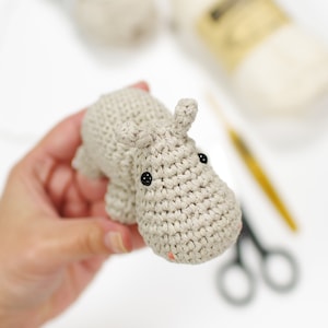 May include: A crocheted gray hippopotamus toy with black eyes. The hippopotamus is being held by a hand.