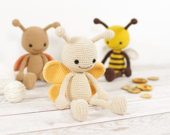 May include: Three handmade crochet insect toys: a butterfly with yellow wings, a bee with black and yellow stripes, and a brown insect. The toys are sitting on a white wooden surface with yarn and buttons.
