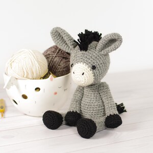 May include: A handmade gray crochet donkey with black hooves and a black mane sits next to a white bowl filled with yarn. The donkey has a white face and ears. Two crochet hooks are visible.