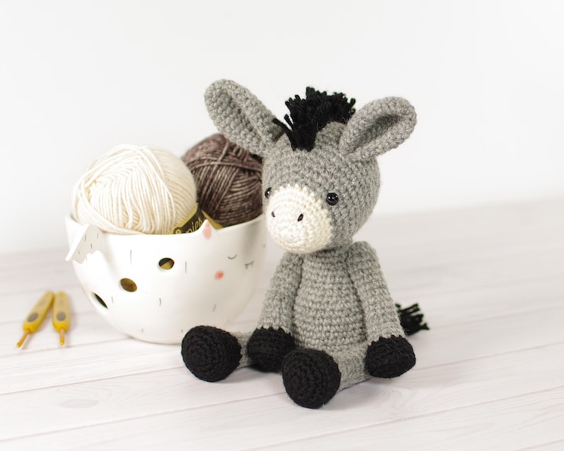 May include: A handmade gray crochet donkey with black hooves and a black mane sits next to a white bowl filled with yarn. The donkey has a white face and ears. Two crochet hooks are visible.