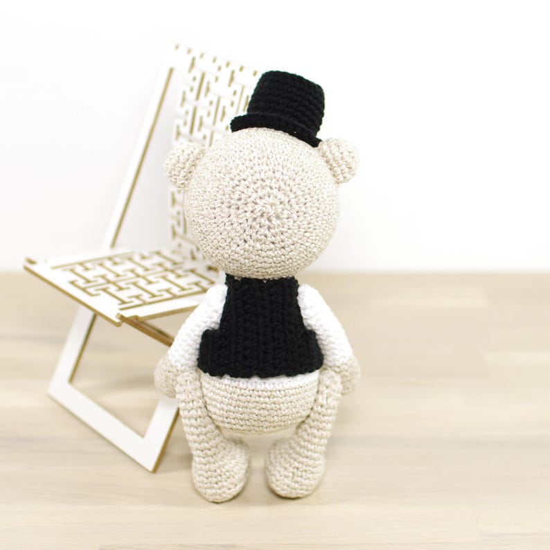 May include: A white crocheted teddy bear wearing a black vest and a black top hat. The bear is standing on a wooden surface.