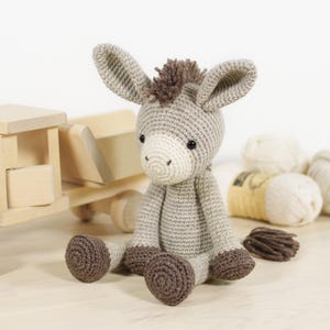 May include: A crocheted donkey soft toy made from light grey yarn, featuring a cream-coloured muzzle, dark brown hooves, and a dark brown mane and tail. The toy has small black safety eyes and long, upright ears, created with a detailed stitch pattern.