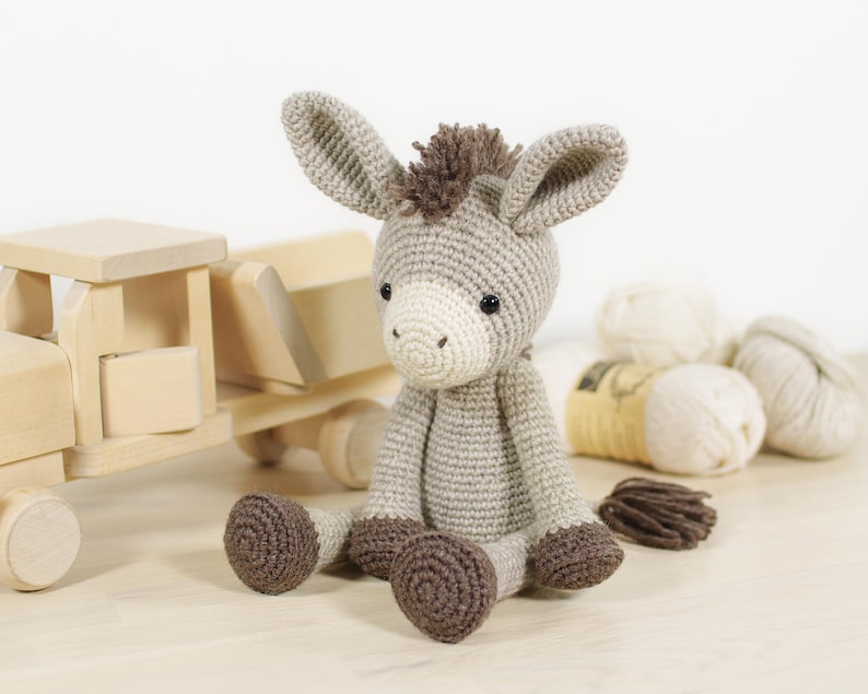 May include: A crocheted donkey soft toy made from light grey yarn, featuring a cream-coloured muzzle, dark brown hooves, and a dark brown mane and tail. The toy has small black safety eyes and long, upright ears, created with a detailed stitch pattern.