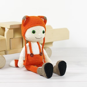 May include: A crocheted fox doll wearing an orange jumpsuit and a fox hat with white accents. The doll has green eyes and a button nose. The doll is sitting on a wooden surface.