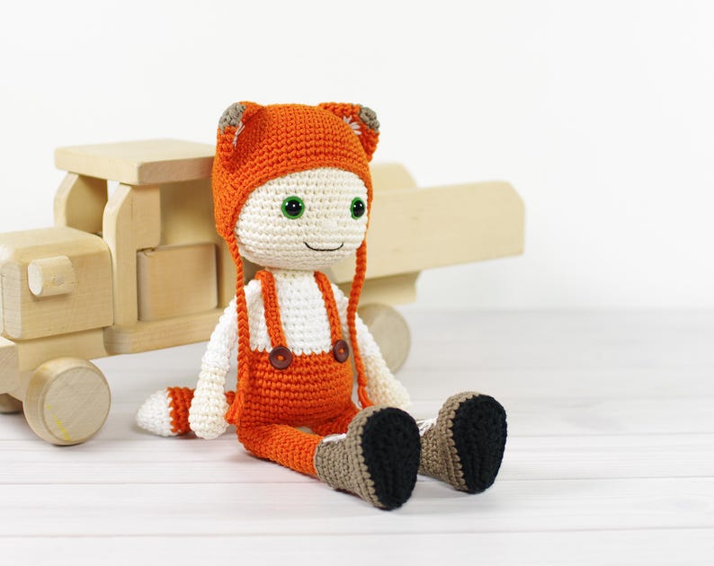 May include: A crocheted fox doll wearing an orange jumpsuit and a fox hat with white accents. The doll has green eyes and a button nose. The doll is sitting on a wooden surface.