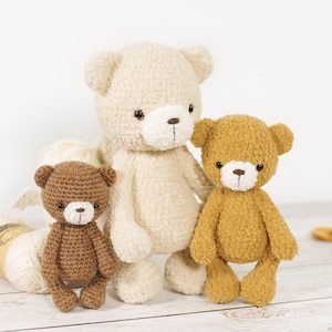 May include: Three handmade crochet teddy bears in varying sizes and shades of brown and cream. The bears have button eyes and stitched noses. A ball of yarn and wooden buttons are in the background, suggesting a craft project.