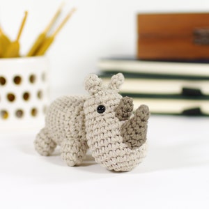 May include: A crocheted rhinoceros toy with a brown horn and black eyes. The toy is made of beige yarn and is sitting on a white surface.