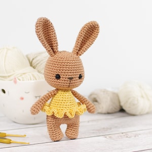 Small caramel brown crocheted bunny in a yellow ballerina skirt, standing in front of a yarn bowl, crochet hooks and balls of yarn