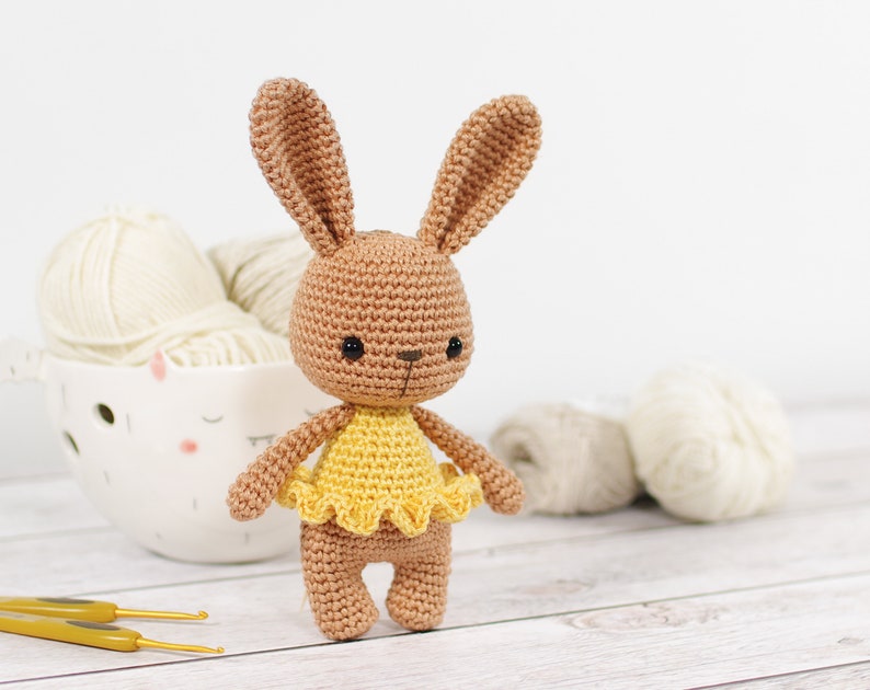 Small caramel brown crocheted bunny in a yellow ballerina skirt, standing in front of a yarn bowl, crochet hooks and balls of yarn