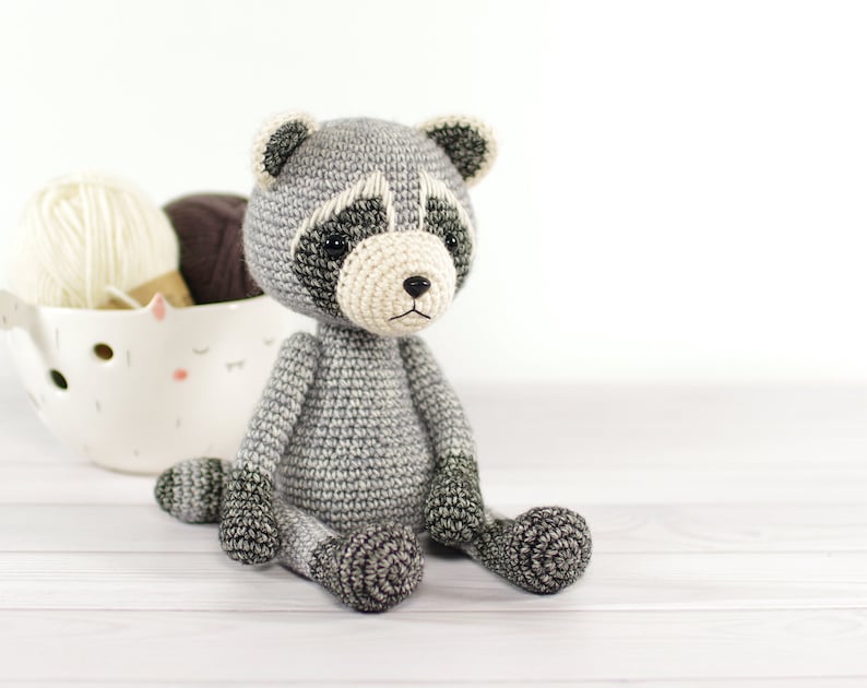 May include: A crocheted raccoon plush toy with gray and black fur, sitting on a white wooden surface. The toy has black eyes, a white nose, and a black mouth.