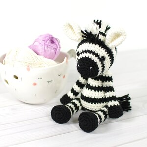K&ouml;nnte beinhalten: Ein schwarz-wei&szlig; gestreiftes geh&auml;keltes Zebra-Spielzeug, das auf einer wei&szlig;en Holzoberfl&auml;che sitzt. Das Zebra hat schwarze Augen und eine schwarze Nase. Hinter dem Zebra befindet sich eine wei&szlig;e Keramikschale mit Garn.