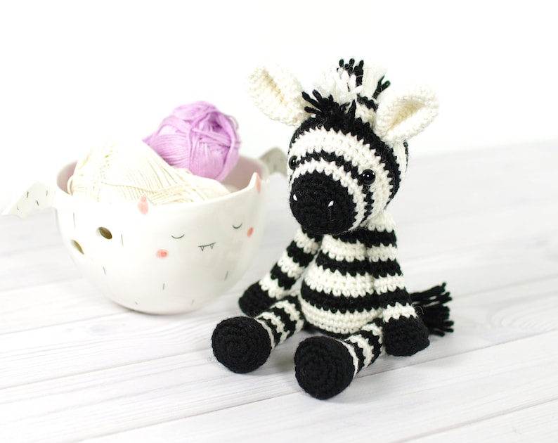 K&ouml;nnte beinhalten: Ein schwarz-wei&szlig; gestreiftes geh&auml;keltes Zebra-Spielzeug, das auf einer wei&szlig;en Holzoberfl&auml;che sitzt. Das Zebra hat schwarze Augen und eine schwarze Nase. Hinter dem Zebra befindet sich eine wei&szlig;e Keramikschale mit Garn.