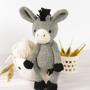 May include: A handmade crochet donkey doll in shades of gray, black, and cream. The donkey has large ears, a black mane, and black hooves. A ball of cream-colored yarn and crochet hooks are in the background.