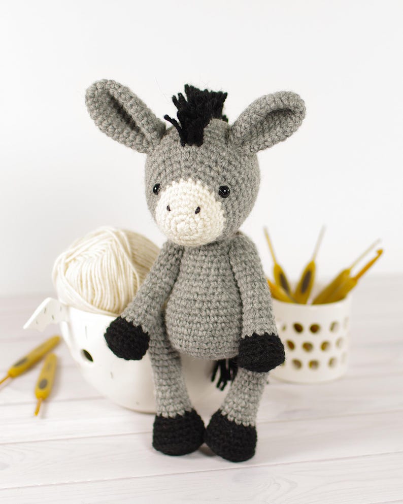 May include: A handmade crochet donkey doll in shades of gray, black, and cream. The donkey has large ears, a black mane, and black hooves. A ball of cream-colored yarn and crochet hooks are in the background.