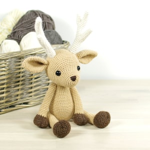 May include: A crocheted fawn toy with brown and white fur, sitting on a wooden surface. The fawn has large brown eyes and a small brown nose.