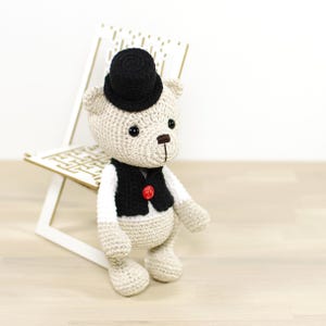 May include: A crocheted teddy bear wearing a black top hat and a black vest with a red button. The bear is sitting on a white wooden chair.