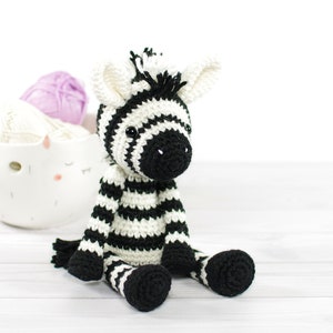 May include: A crocheted zebra toy with black and white stripes. The zebra is sitting on a white wooden surface.