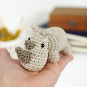May include: A small crocheted rhinoceros toy in beige and brown with black eyes. The toy is being held in a person's hand.
