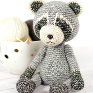 May include: A crocheted gray raccoon plush toy with black eyes and a white nose. The toy is sitting on a white wooden surface.