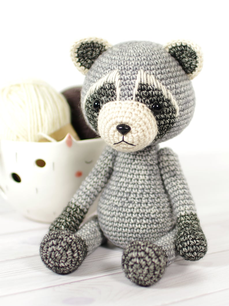 May include: A crocheted gray raccoon plush toy with black eyes and a white nose. The toy is sitting on a white wooden surface.