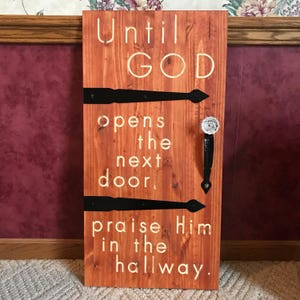 May include: Wooden sign with the text "Until GOD opens the next door, praise Him in the hallway." The sign is stained a reddish-brown color and features black hinges and a decorative handle.