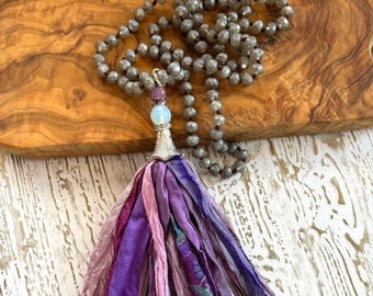 May include: A purple and pink silk tassel necklace with a silver charm and a light blue gemstone. The necklace is made of gray beads.