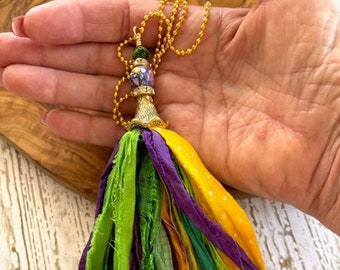 May include: A colorful tassel necklace with a gold chain and a purple and green bead. The tassel is made of green, purple, yellow, and orange fabric.