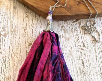 May include: A long silver chain necklace with a large tassel pendant. The tassel is made of layered fabric in shades of pink and purple. The pendant has a silver cap with a beaded accent and a small purple bead.