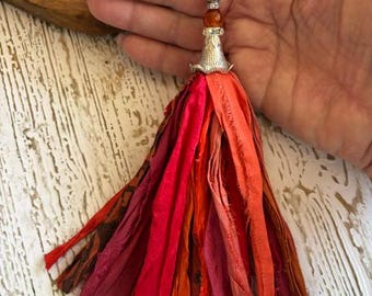 May include: A tassel pendant featuring long strips of fabric in shades of red, orange, and burgundy. The tassel is topped with a silver-toned metal cap, a small orange bead, and a speckled brown bead, all suspended from a silver-toned ball chain.