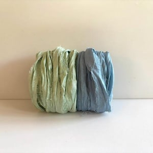 May include: Two spools of crinkled silk ribbon, one in a light sage green and the other in a soft dusty blue. Each ribbon features a textured, frayed edge appearance with a matte finish, wound tightly into a cylindrical shape side by side.