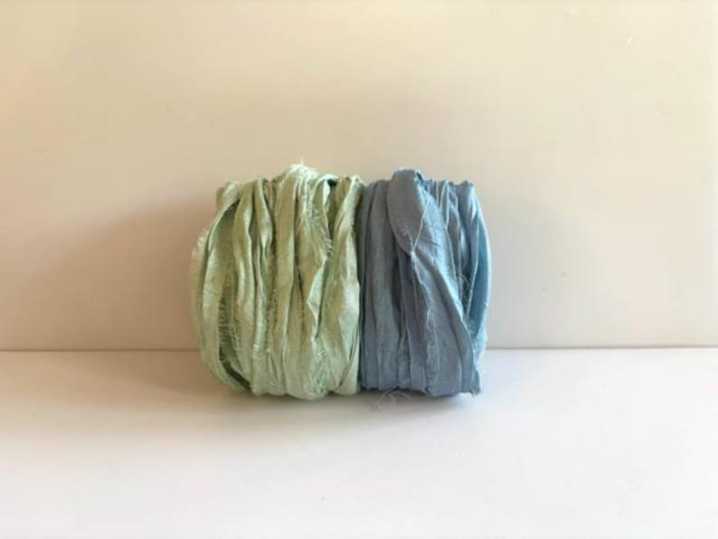 May include: Two spools of crinkled silk ribbon, one in a light sage green and the other in a soft dusty blue. Each ribbon features a textured, frayed edge appearance with a matte finish, wound tightly into a cylindrical shape side by side.