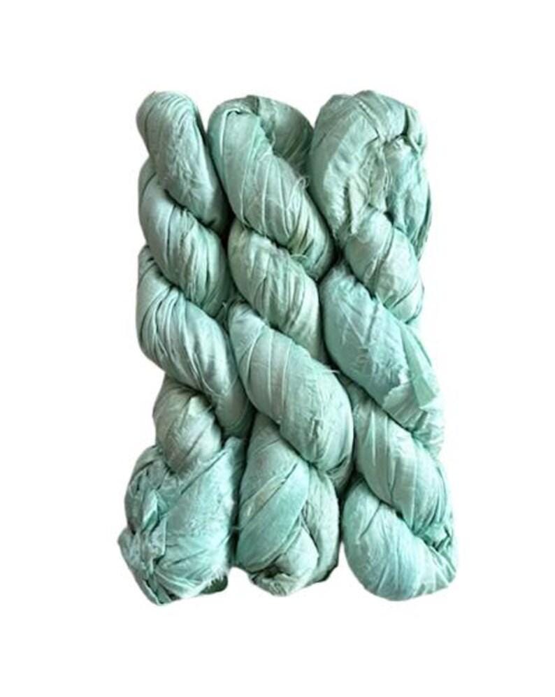 May include: A bundle of light mint green silk ribbon yarn featuring a textured, crinkled appearance. The material is wound into a series of interconnected loops, displaying a soft, matte finish with visible frayed edges along the length of the fabric.