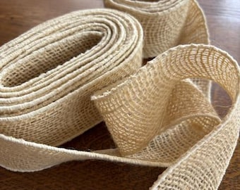 Puede incluir: Primer plano de cinta de arpillera de color natural, enrollada y desenrollada. La cinta tiene una textura tejida y está hecha de un material de color beige claro. La cinta está sobre una superficie de madera marrón oscuro.