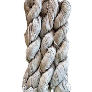 May include: A bundle of light taupe silk ribbon yarn featuring a crinkled, textured surface. The material is wound into a long, thick skein with visible frayed edges and a soft, matte sheen, displaying a consistent neutral color throughout.