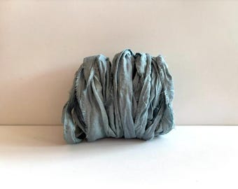 May include: A bundle of dusty blue silk ribbon with a frayed, raw edge texture. The fabric is loosely coiled and bunched together, displaying a matte finish and a soft, wrinkled appearance throughout the length of the material.