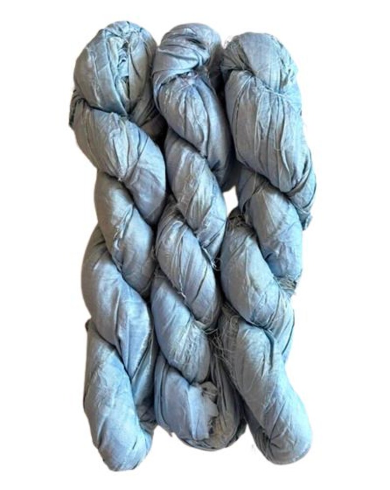May include: Three skeins of light blue ribbon yarn arranged vertically. The yarn has a crinkled, matte texture with visible frayed edges along the strands. The color is a consistent pale sky blue throughout the entire length of the material.