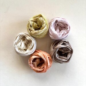 May include: Six spools of colorful, textured ribbon arranged in a circle. The ribbons are in shades of cream, pale pink, peach, olive green, and brown. The ribbons appear to be made of a soft, natural material.