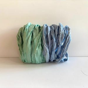 May include: A bundle of fabric ribbons in shades of seafoam green and blue. The ribbons are tightly gathered, creating a textured, layered appearance. The ribbons are arranged side by side, with the green on the left and the blue on the right.