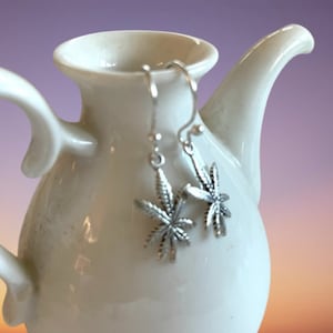 Könnte beinhalten: Silberne Ohrringe mit einem Marihuana-Blatt-Design, die an einer weißen Keramik-Teekanne hängen.