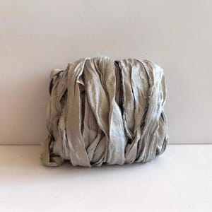 May include: A bundle of light gray silk ribbon with a crinkled, textured surface and frayed edges. The ribbon is wound into a compact, rectangular shape, showcasing a soft, matte finish and a consistent neutral color throughout the entire length.